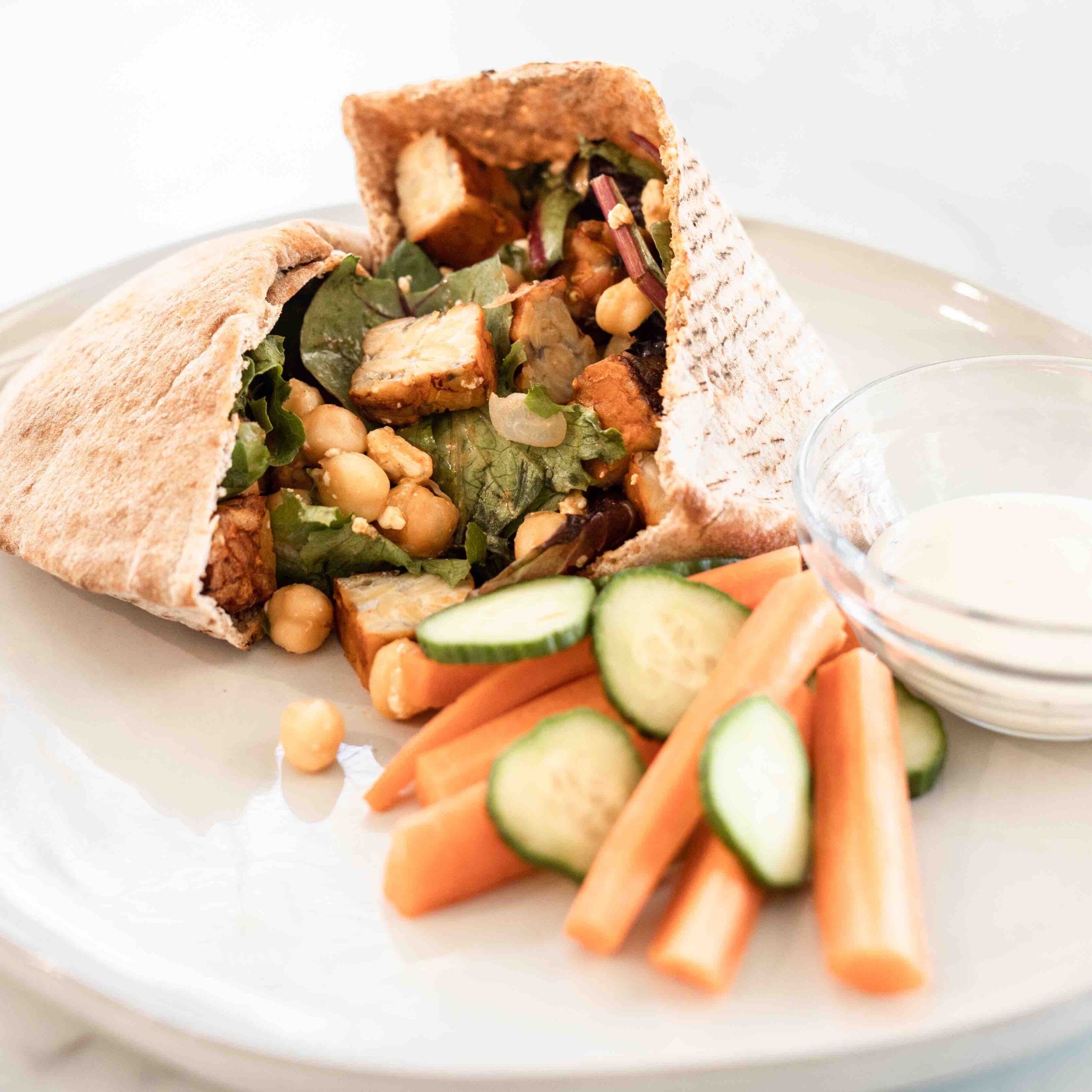 A zoomed-in view of a stuffed whole wheat pita pocket, showing crispy buffalo tempeh, chickpeas, and mixed greens spilling out. The plate also features fresh cucumber slices, carrot sticks, and a side of ranch dressing.