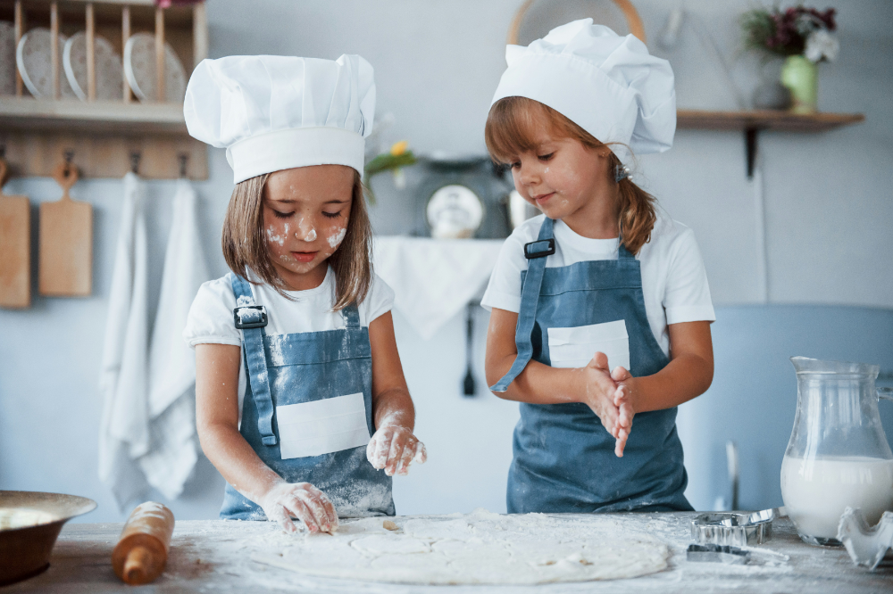 Two kids in blue aprons with chef hats making pizza dough